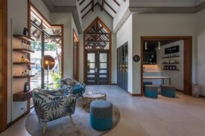 Moderní prosluněný salonek se vzorovanými křesly, kulatými otomany a&nbsp;malým stolkem. Velká okna a&nbsp;dřevěné akcenty vytvářejí příjemnou, vzdušnou atmosféru. V&nbsp;pozadí jsou vidět police s&nbsp;výrobky a&nbsp;recepce.