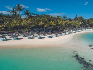 Tropická pláž s&nbsp;průzračnou tyrkysovou vodou, bílým pískem, řadami modrých lehátek a&nbsp;slaměných slunečníků lemovaných vysokými palmami a&nbsp;letoviskem za&nbsp;nimi pod jasně modrou oblohou.