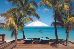 Na dřevěné terase mezi palmami stojí dvě lehátka a&nbsp;bílý slunečník s&nbsp;výhledem na&nbsp;klidné tyrkysové moře a&nbsp;malou loďku plující v&nbsp;dálce pod jasně modrou oblohou.