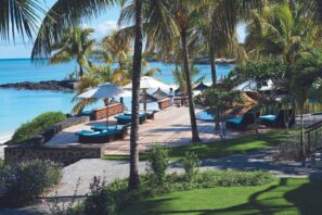 Tropický plážový resort s&nbsp;dřevěnou terasou, lehátky a&nbsp;bílými slunečníky s&nbsp;výhledem na&nbsp;tyrkysovou vodu, obklopený palmami a&nbsp;bujnou zelení pod slunečnou oblohou.