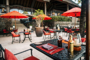 Venkovní terasa restaurace s&nbsp;černými stoly, červenými polstrovanými židlemi a&nbsp;červenými slunečníky. Stoly jsou prostřené sklenicemi, ubrousky a&nbsp;lahvemi. V&nbsp;pozadí jsou vidět kamenné zdi a&nbsp;schody a&nbsp;zeleň.