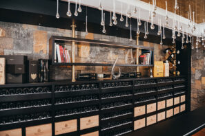 Moderní vinný sklep s&nbsp;policemi s&nbsp;lahvemi vína, dřevěnými bednami, knihami, skleněnými karafami a&nbsp;dekorativními svítidly zavěšenými u&nbsp;stropu na&nbsp;kamenné zdi.