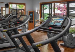 Moderní posilovna s&nbsp;běžeckými pásy se zabudovanými obrazovkami, velkými okny, dřevěnými podlahami a&nbsp;výhledem do&nbsp;svěží venkovní zahrady s&nbsp;terasovým nábytkem a&nbsp;tropickými rostlinami.