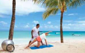 Žena cvičí jógu v&nbsp;bočním protažení na&nbsp;podložce mezi dvěma palmami na&nbsp;písečné pláži, zatímco jí muž pomáhá. V&nbsp;pozadí pod jasně modrou oblohou je cvičební nářadí a&nbsp;klidné tyrkysové moře.