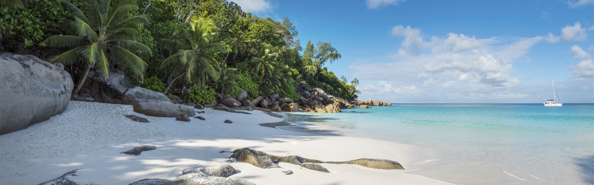Tropická pláž s&nbsp;bílým pískem, palmami a&nbsp;velkými skalami podél pobřeží. Břehu se dotýká průzračná tyrkysová voda a&nbsp;na klidném moři pod jasně modrou oblohou s&nbsp;roztroušenými mraky pluje plachetnice.