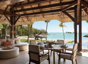 Z jídelny a&nbsp;lounge pod širým nebem s&nbsp;doškovou střechou je výhled na&nbsp;tropickou pláž, palmy a&nbsp;tyrkysový oceán. Kulatý stůl je prostřen skleněným nádobím a&nbsp;prostor obklopují polstrovaná sedadla.