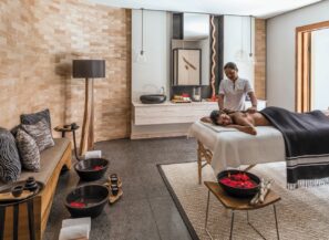 V moderní, klidné lázeňské místnosti s&nbsp;dřevěnými akcenty, polstrovanou lavicí, miskami s&nbsp;vodou s&nbsp;plovoucími červenými květy a&nbsp;jemným osvětlením se ženě dostává masáže zad od terapeuta.