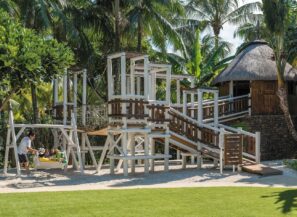 Moderní venkovní hřiště s&nbsp;dřevěnými prolézačkami, skluzavkami a&nbsp;houpačkou, obklopené palmami a&nbsp;zelení. Dospělý tlačí děti na&nbsp;houpačce u&nbsp;dřevěné budovy se slaměnou střechou.