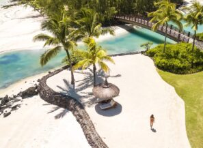 Žena se prochází po&nbsp;písečné pláži u&nbsp;slaměného slunečníku a&nbsp;palem, v&nbsp;pozadí je kamenná zeď a&nbsp;tyrkysová laguna, přes kterou vede dřevěný most.