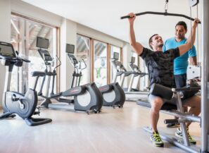 Muž cvičí v&nbsp;posilovně na&nbsp;stroji na&nbsp;shyby, zatímco mu jiný muž, pravděpodobně trenér, pomáhá. V&nbsp;pozadí u&nbsp;velkých oken je vidět několik běžeckých pásů a&nbsp;stacionárních kol.