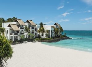 Bílé budovy resortu na&nbsp;pláži s&nbsp;doškovými střechami jsou obklopené palmami, s&nbsp;výhledem na&nbsp;tyrkysový oceán a&nbsp;nedotčenou písečnou pláž pod slunečně modrou oblohou.