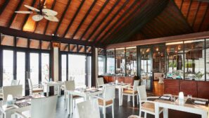 Moderní restaurace s&nbsp;bílými stoly a&nbsp;židlemi, vysokým dřevěným stropem s&nbsp;odhalenými trámy, velkými okny s&nbsp;výhledem ven a&nbsp;barovým pultem se skleněnými stěnami v&nbsp;pozadí.