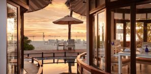 Pohled zevnitř restaurace na&nbsp;pláži ukazuje stoly a&nbsp;židle na&nbsp;písku, slunečník, palmy a&nbsp;klidný oceán při západu slunce s&nbsp;teplým zlatým světlem odrážejícím se od skla a&nbsp;vody.