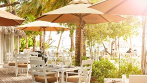 Venkovní restaurace s&nbsp;bílými stoly a&nbsp;židlemi pod velkými béžovými slunečníky, obklopená zelenými rostlinami a&nbsp;palmami, s&nbsp;proudícím slunečním světlem a&nbsp;lidmi stolujícími v&nbsp;pozadí u&nbsp;vody.