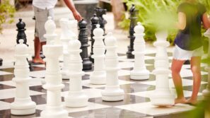 Na velké venkovní šachovnici stojí dvě osoby a&nbsp;pohybují velkými bílými šachovými figurkami. Černé a&nbsp;bílé figurky jsou rozesety po&nbsp;šachovnici a&nbsp;v pozadí je vidět zeleň.