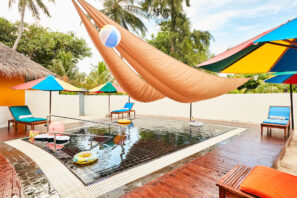 Malý venkovní bazén s&nbsp;barevnými slunečníky, lehátky a&nbsp;plováky. Nad ním je zavěšena velká opálená látková stříška a&nbsp;v pozadí jsou vidět palmy a&nbsp;zeleň. Nad bazénem visí plážový míč.