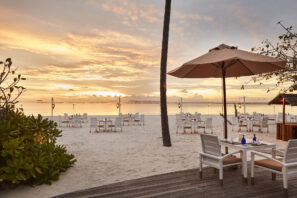 Venkovní restaurace na&nbsp;pláži s&nbsp;bílými stoly a&nbsp;židlemi na&nbsp;písku s&nbsp;výhledem na&nbsp;oceán při západu slunce. Jeden stůl zastiňuje slunečník a&nbsp;obloha je plná teplých oranžových a&nbsp;žlutých odstínů.