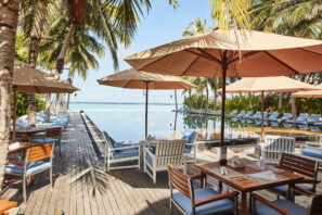 Venkovní jídelna s&nbsp;dřevěnými stoly a&nbsp;židlemi pod velkými slunečníky vedle klidného nekonečného bazénu obklopeného palmami a&nbsp;lehátky s&nbsp;výhledem na&nbsp;modrou oblohu a&nbsp;oceán.