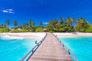 Dřevěná promenáda vede přes tyrkysovou vodu na&nbsp;tropickou pláž lemovanou palmami a&nbsp;bujnou zelení pod jasně modrou oblohou.