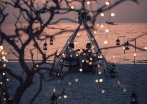 Pár večeří pod bílým baldachýnem na&nbsp;písečné pláži při západu slunce, obklopen lampiony a&nbsp;pohádkovými světly, s&nbsp;oceánem a&nbsp;vzdáleným hydroplánem v&nbsp;pozadí.