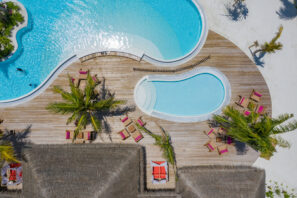 Letecký pohled na&nbsp;tropické letovisko se zakřiveným bazénem, dřevěnou terasou, lehátky s&nbsp;červenými polštáři, palmami a&nbsp;chatkami s&nbsp;doškovou střechou na&nbsp;bílém písku.