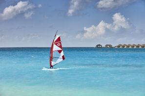 Osoba na&nbsp;windsurfingu na&nbsp;jasně tyrkysové vodě pod částečně zataženou oblohou, v&nbsp;dálce jsou vidět bungalovy nad vodou.