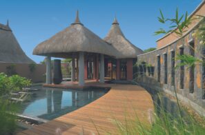 Tropická vila s&nbsp;doškovými střechami, dřevěným chodníkem a&nbsp;malým bazénem obklopená bujnou zelení a&nbsp;kamennými zdmi pod jasně modrou oblohou.