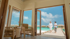 Pohled ze světlého otevřeného pokoje s proutěným nábytkem a vázou na dřevěnou promenádu nad tyrkysovou vodou, po které se procházejí dva lidé v bílém pod slunečně modrou oblohou s bungalovy nad vodou v pozadí.