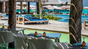 Venkovní restaurace s proutěnými židlemi a prostřenými stoly má výhled na bazén a chatky s doškovou střechou u tyrkysové vody; sluneční světlo a stíny vytvářejí uvolněnou tropickou atmosféru.