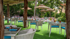 Venkovní restaurace s proutěnými židlemi, skleněnými stoly s ubrousky a sklenicemi, pod dřevěnou pergolou. Slunečníky a další místa k sezení jsou k vidění na zelené umělé trávě, obklopené bujnými stromy a rostlinami.