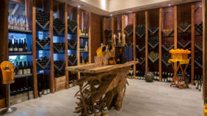 Vinný sklípek s dřevěnými regály naplněnými lahvemi vína, rustikální dřevěný stůl uprostřed se sklenicemi a lahvemi šampaňského a koš pomerančů na stojanu vpravo. Teplé osvětlení vytváří útulnou atmosféru.