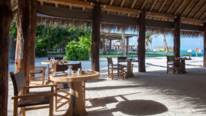 Restaurace pod širým nebem na pláži s dřevěnými stoly a židlemi na písečné podlaze pod doškovou střechou s výhledem do svěží zahrady a na průzračně modrý oceán s palmami v pozadí.