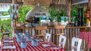 Rustikální venkovní restaurace nabízí stoly s červenobílými kostkovanými ubrusy, talíře, ubrousky a modré sklenice. V pozadí se nachází pec na pizzu vytápěná dřevem a dřevěný bar obklopený zelení.