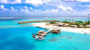 Letecký pohled na tropický ostrov s bungalovy nad vodou, průzračnou tyrkysovou vodou, přístavištěm s hydroplánem a bílou písečnou pláží obklopenou palmami pod jasně modrou oblohou s mraky.