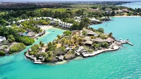 Letecký pohled na tropický ostrov s bujnou zelení, palmami, bílými budovami a chatkami s doškovými střechami, obklopený tyrkysovou vodou a živým pobřežím.