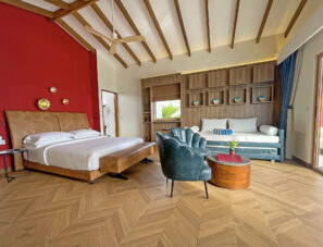 Moderní hotelový pokoj s&nbsp;manželskou postelí, modrými křesly, denním lůžkem, dřevěnou podlahou, výraznou červenou stěnou, stropním ventilátorem a&nbsp;velkými okny propouštějícími přirozené světlo.