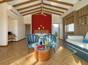 Moderní ložnice s&nbsp;dřevěnou podlahou, velkou postelí na&nbsp;červené stěně, dvěma modrými křesly kolem malého kulatého stolku, modrou pohovkou a&nbsp;psacím stolem s&nbsp;židlí v&nbsp;rohu. Odkryté stropní trámy a&nbsp;velká okna dodávají světlo.