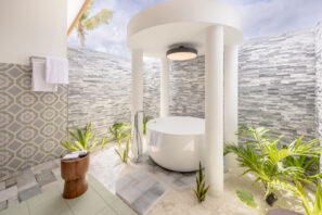 V moderní venkovní koupelně je kulatá bílá vana pod kruhovým baldachýnem, obklopená šedými kamennými stěnami, zelenými tropickými rostlinami a&nbsp;vzorovanými dlaždicemi, do&nbsp;které shora proudí sluneční světlo.