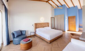 Světlý, moderní hotelový pokoj s&nbsp;manželskou postelí, opáleným čelem postele, dřevěnou podlahou, stěnou s&nbsp;modrým akcentem, křeslem, otomanem s&nbsp;červenou knihou, stropním ventilátorem a&nbsp;přirozeným světlem z&nbsp;velkých oken se závěsy.