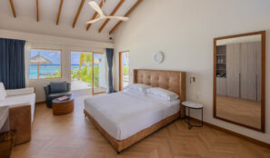 Světlá moderní ložnice s&nbsp;velkou postelí, dřevěnou podlahou, pohovkou a&nbsp;zrcadlem. Okna od podlahy ke stropu odhalují terasu, svěží zeleň a&nbsp;výhled na&nbsp;oceán s&nbsp;chatkou nad vodou.