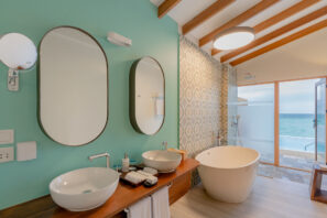 Moderní koupelna s&nbsp;tyrkysovou stěnou, dvěma oválnými zrcadly nad dvojitým umyvadlem, volně stojící bílou vanou, dřevěnými trámy na&nbsp;stropě a&nbsp;velkým oknem s&nbsp;výhledem na&nbsp;oceán.