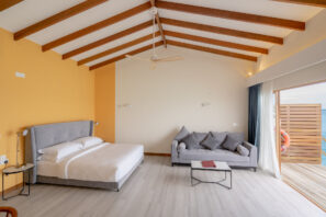 Moderní vzdušná ložnice s&nbsp;klenutým stropem a&nbsp;dřevěnými trámy je vybavena šedou postelí, šedou pohovkou s&nbsp;polštáři, malým konferenčním stolkem a&nbsp;skleněnými dveřmi na&nbsp;balkon, ze kterého je vidět voda a&nbsp;záchranný kruh.