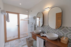 Moderní koupelna se dvěma umyvadly na&nbsp;dřevěném pultu, oválnými zrcadly, vzorovanými obklady, skleněným sprchovým koutem, dveřmi z&nbsp;matného skla, ručníky a&nbsp;proutěným košem na&nbsp;prádlo na&nbsp;podlaze.