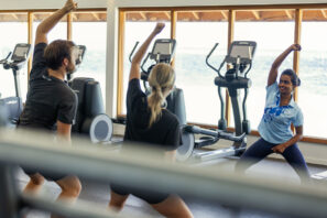 Tři lidé ve&nbsp;sportovním oblečení se účastní skupinové lekce fitness v&nbsp;tělocvičně, protahují se a&nbsp;zvedají ruce, v&nbsp;pozadí jsou cvičební stroje a&nbsp;velká okna s&nbsp;výhledem na&nbsp;vodu.