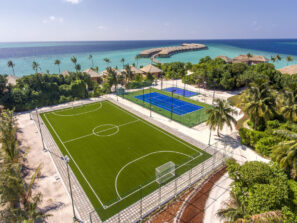 Letecký pohled na&nbsp;tropické letovisko se zeleným fotbalovým hřištěm a&nbsp;modrým tenisovým kurtem, obklopené palmami, vedle tyrkysové vody oceánu a&nbsp;bungalovů nad vodou pod slunečnou jasnou oblohou.
