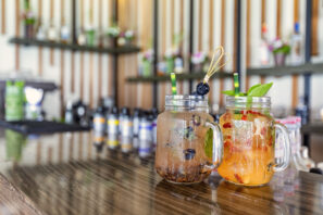 Na lesklém dřevěném barovém pultu stojí dvě zavařovací sklenice naplněné barevnými koktejly. Jeden nápoj je s&nbsp;borůvkami a&nbsp;brčkem, druhý s&nbsp;plátky citrusů a&nbsp;bylinkami. V&nbsp;pozadí jsou láhve a&nbsp;rozmazané police.