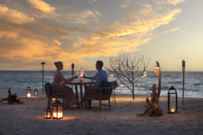 Pár sedí u&nbsp;stolu na&nbsp;písečné pláži při západu slunce, obklopen lucernami, svíčkami a&nbsp;větví stromu ozdobenou závěsnými světly, s&nbsp;oceánem a&nbsp;barevnou oblohou v&nbsp;pozadí.