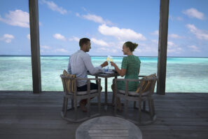 Muž a&nbsp;žena sedí u&nbsp;stolu na&nbsp;terase nad průzračně modrou vodou oceánu a&nbsp;připíjejí si nápoji pod slunečnou oblohou s&nbsp;malebným výhledem na&nbsp;obzor a&nbsp;jemné mraky.