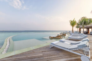 Luxusní nekonečný bazén s&nbsp;lehátky má výhled na&nbsp;klidný modrý oceán. U&nbsp;bazénu jsou dřevěné paluby a&nbsp;stín poskytují palmy. Obloha je jasná a&nbsp;naznačuje klidné a&nbsp;relaxační tropické prostředí.
