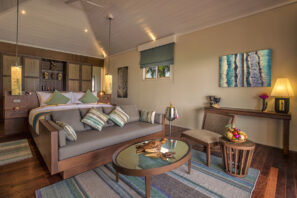 Útulná stylová ložnice s&nbsp;velkou postelí, šedou pohovkou a&nbsp;dřevěným nábytkem. Pokoj má měkké osvětlení, zdobí ho moderní umění, pruhované koberce a&nbsp;na stole je koš s&nbsp;ovocem. Malé okno propouští přirozené světlo.